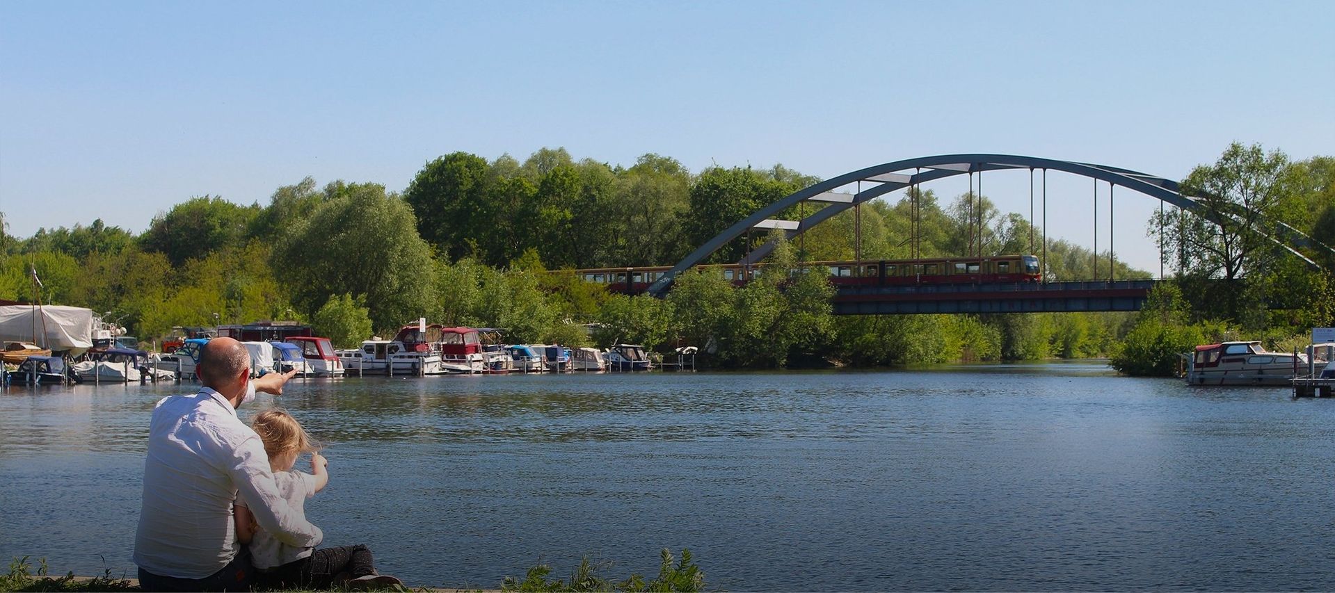 Vater und Kind sitzen an der Spree und beobachten eine Bahn, die über eine Brücke fährt